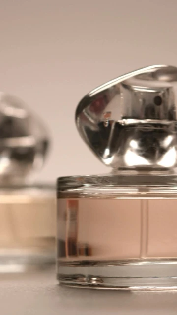 Two perfume bottles with clear glass and silver caps are displayed close-up against a soft, neutral background in vertical format.