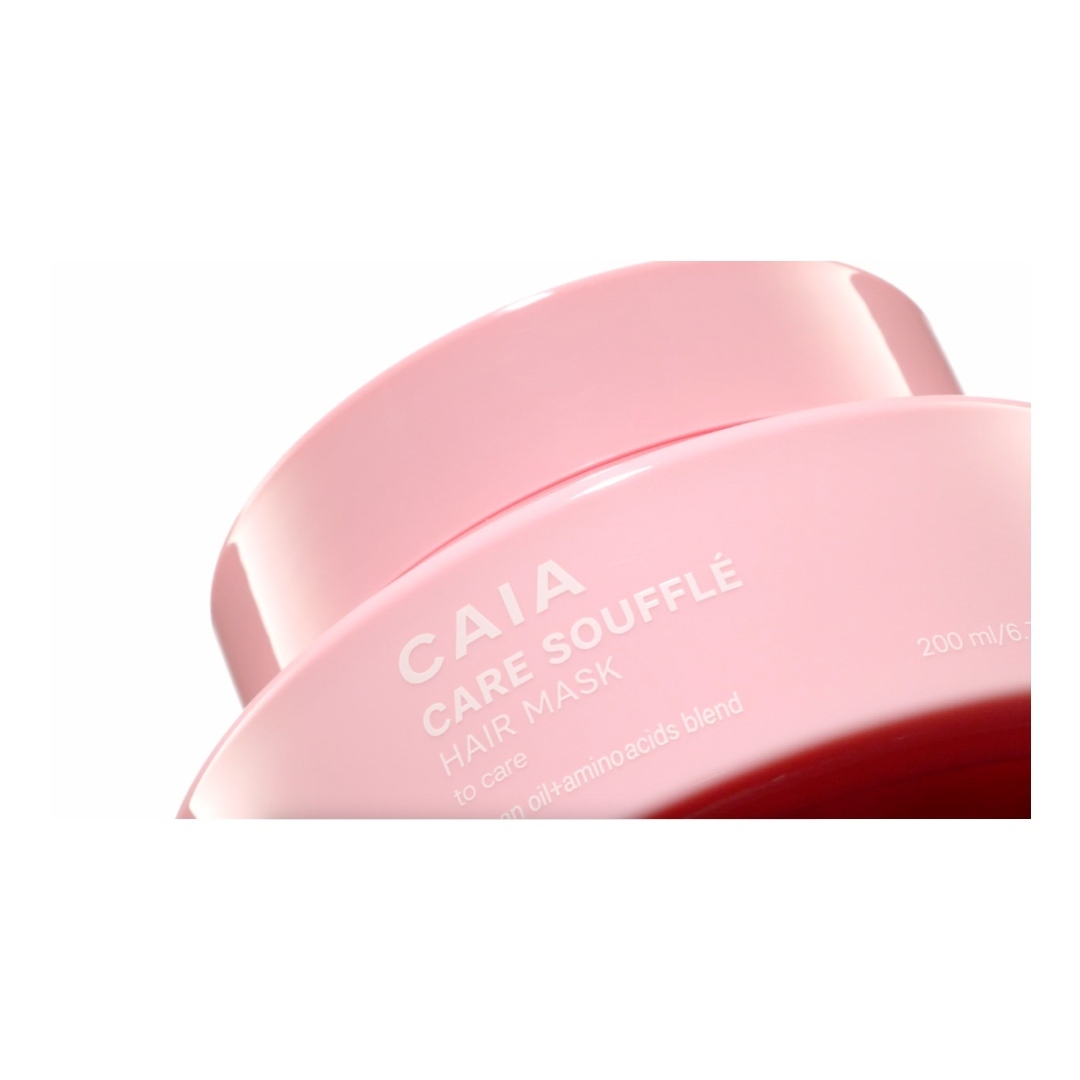 Close-up of a pink hair mask container with 'CAIA Care Soufflé Hair Mask' text, against a bright white backdrop, product photography.