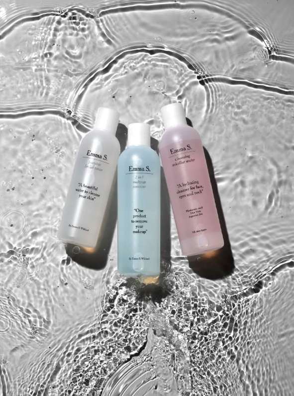 Three colorful Emma S. cleansing water bottles arranged in a line and photographed under rippling water creating abstract patterns.