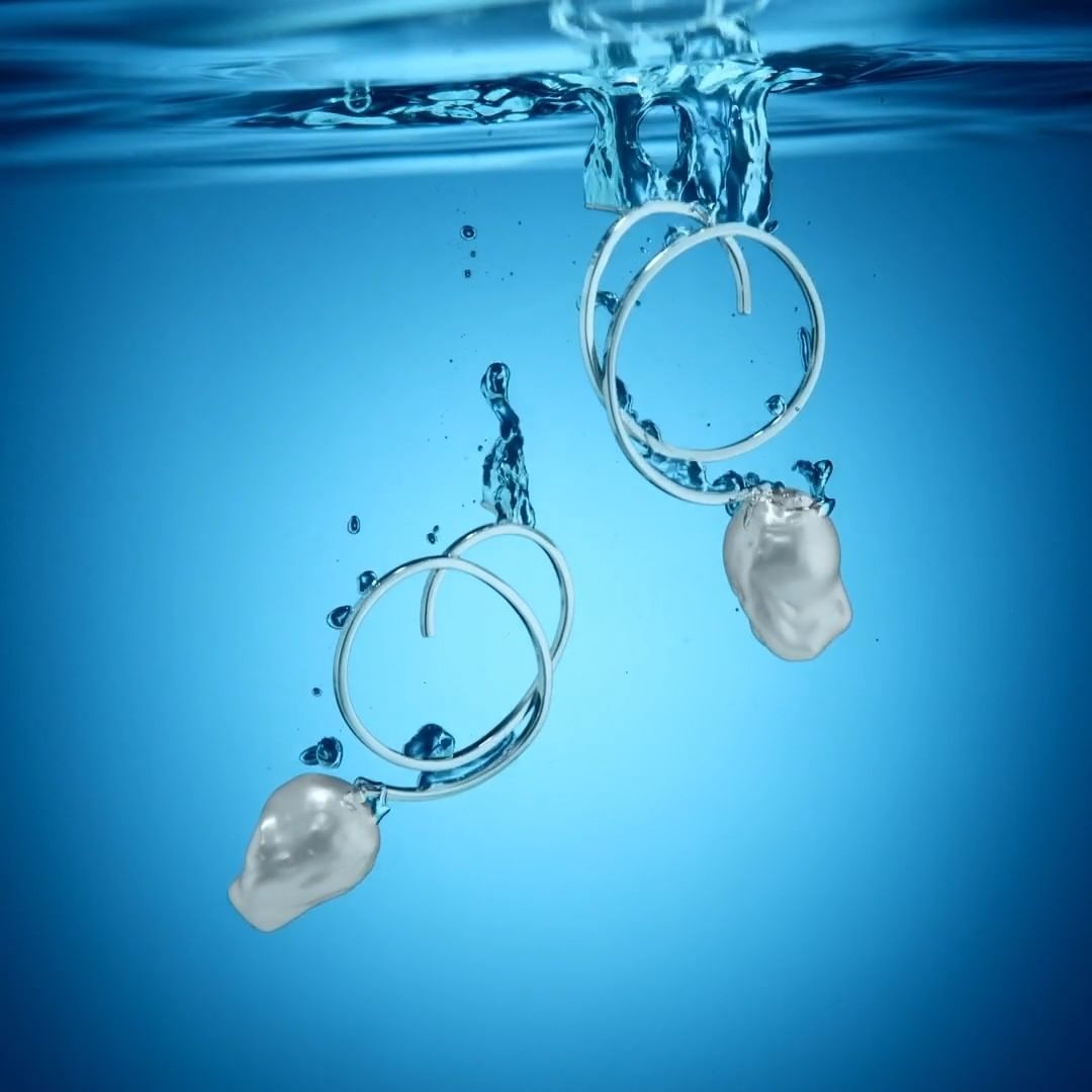 Two earrings with silver loops and white baroque pearls are submerged in clear blue water, creating a splash effect.