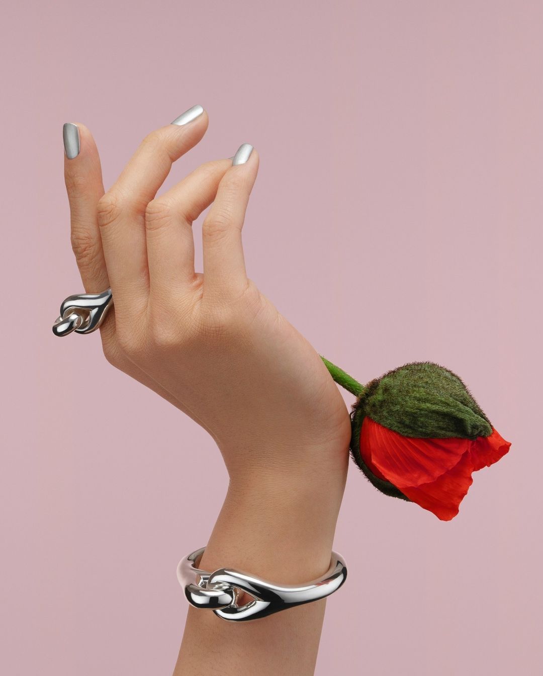 Hand showcases silver ring and bangle with a red poppy on a pink background, metallic nail polish for a beauty or fashion shot.