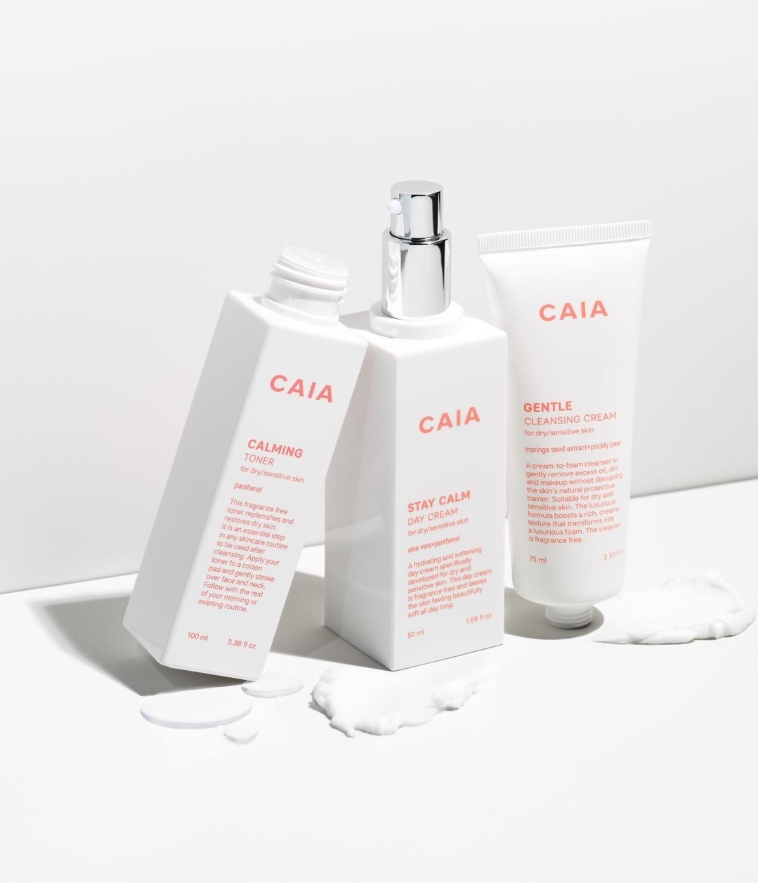 Still life photo featuring a collection of three CAIA Cosmetics skincare products in white packaging, arranged on a white surface with cream.