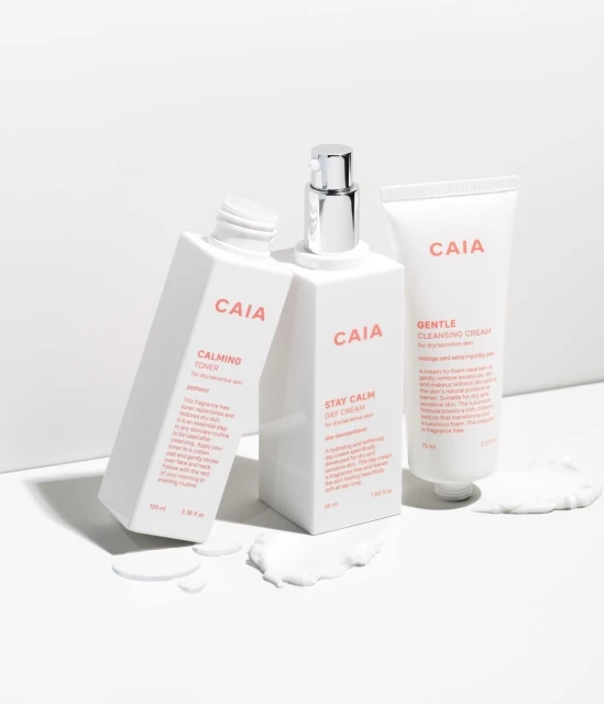 Still life photo featuring a collection of three CAIA Cosmetics skincare products in white packaging, arranged on a white surface with cream.