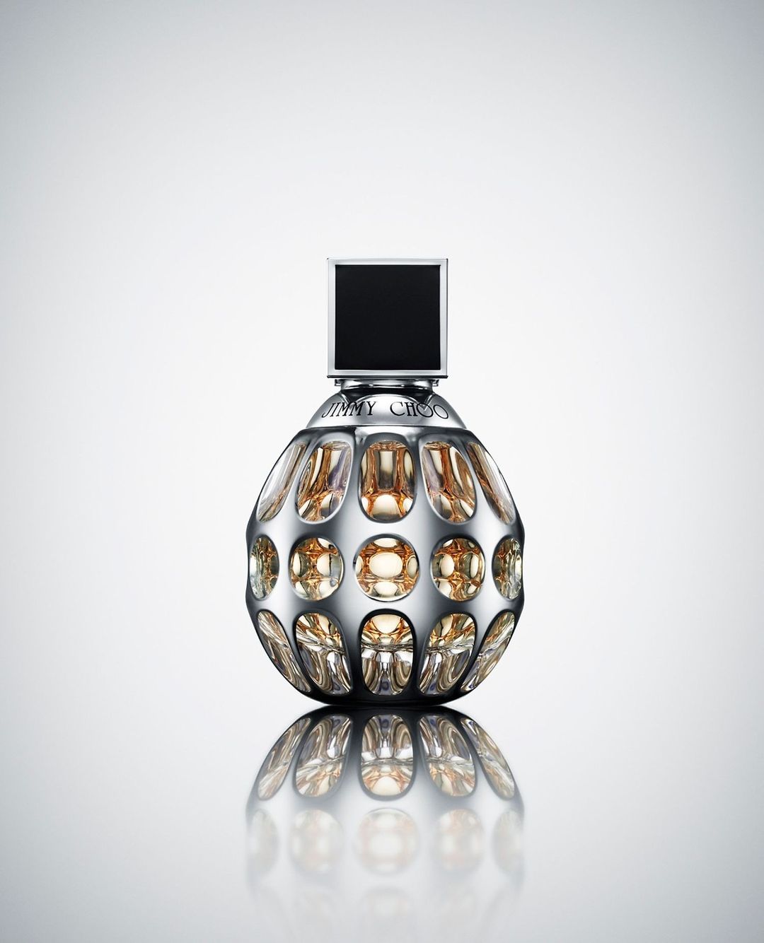 Shiny perfume bottle with a silver spherical body, black square top, and gold liquid inside on a reflective white surface.
