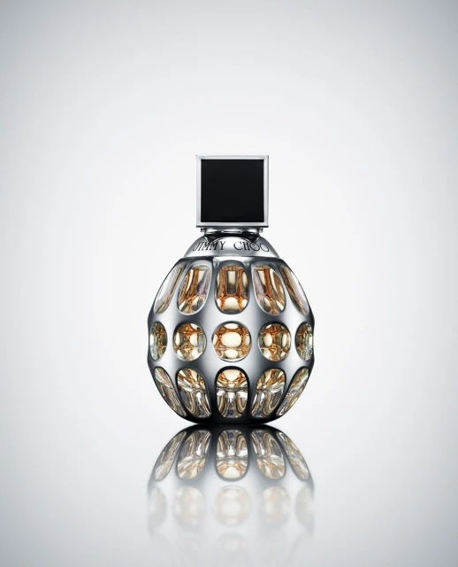 Shiny perfume bottle with a silver spherical body, black square top, and gold liquid inside on a reflective white surface.