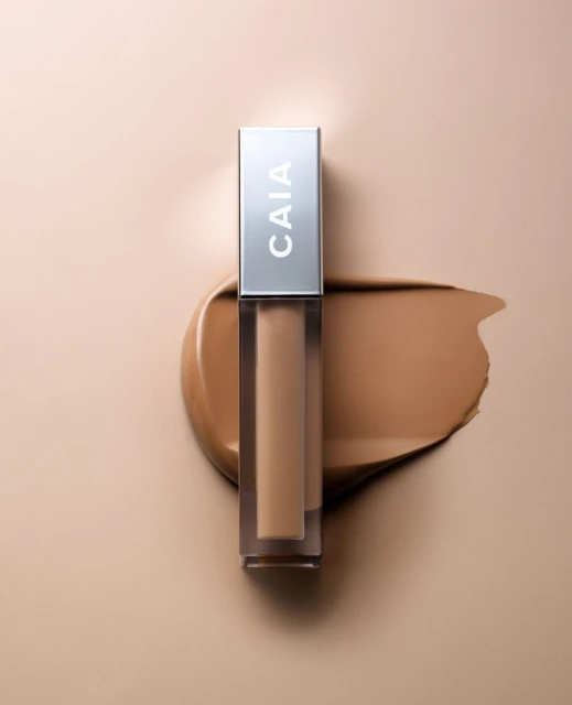 Studio shot of CAIA Cosmetics foundation with neutral tan color swatch on beige background. The product is a modern beauty concept.