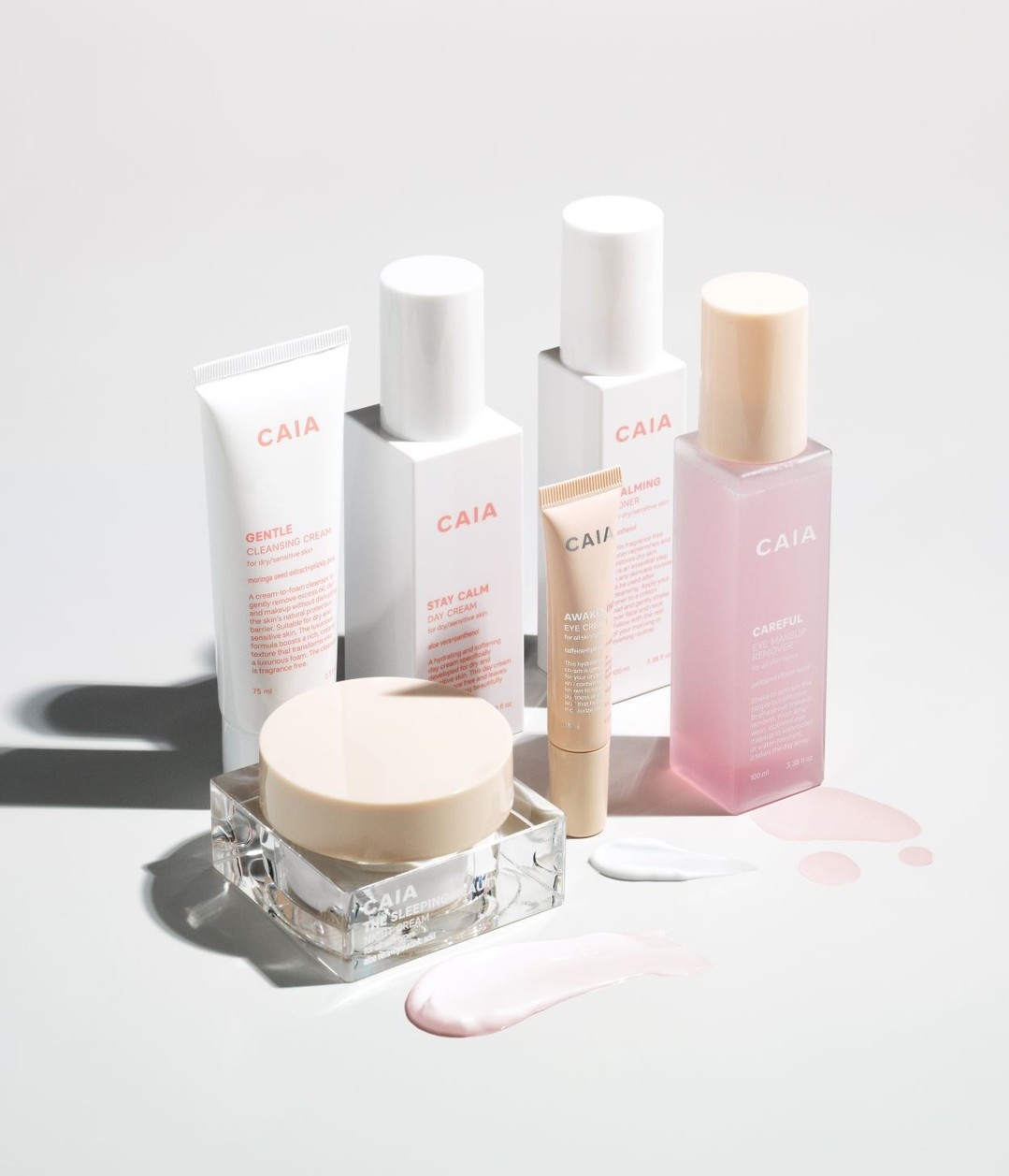 Still life of CAIA Cosmetics skincare products in white packaging, jars, tubes, and bottles on a white surface with product swatches.