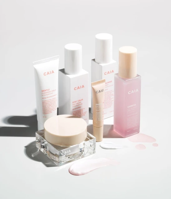 Still life of CAIA Cosmetics skincare products in white packaging, jars, tubes, and bottles on a white surface with product swatches.