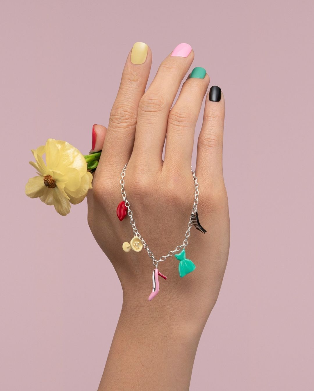 Close-up of a hand holding a yellow flower and adorned with a charm bracelet featuring colorful charms against a light purple backdrop.