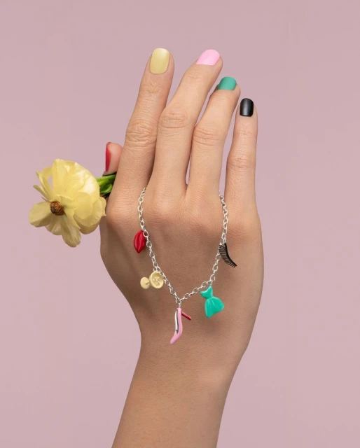 Close-up of a hand holding a yellow flower and adorned with a charm bracelet featuring colorful charms against a light purple backdrop.