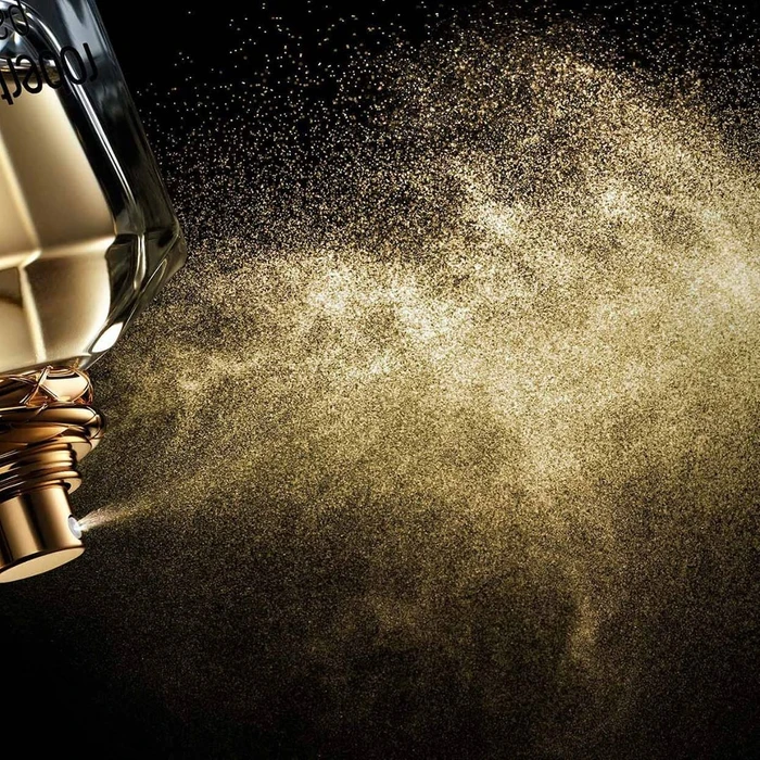 Close-up of a luxury perfume bottle spraying fine golden mist and glitter against a dark background, captured by Daniel Lindh.