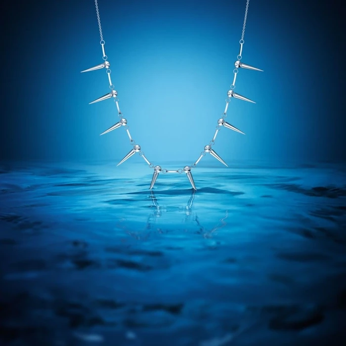 Silver spike necklace suspended over rippling deep blue water reflecting light in a studio setting.