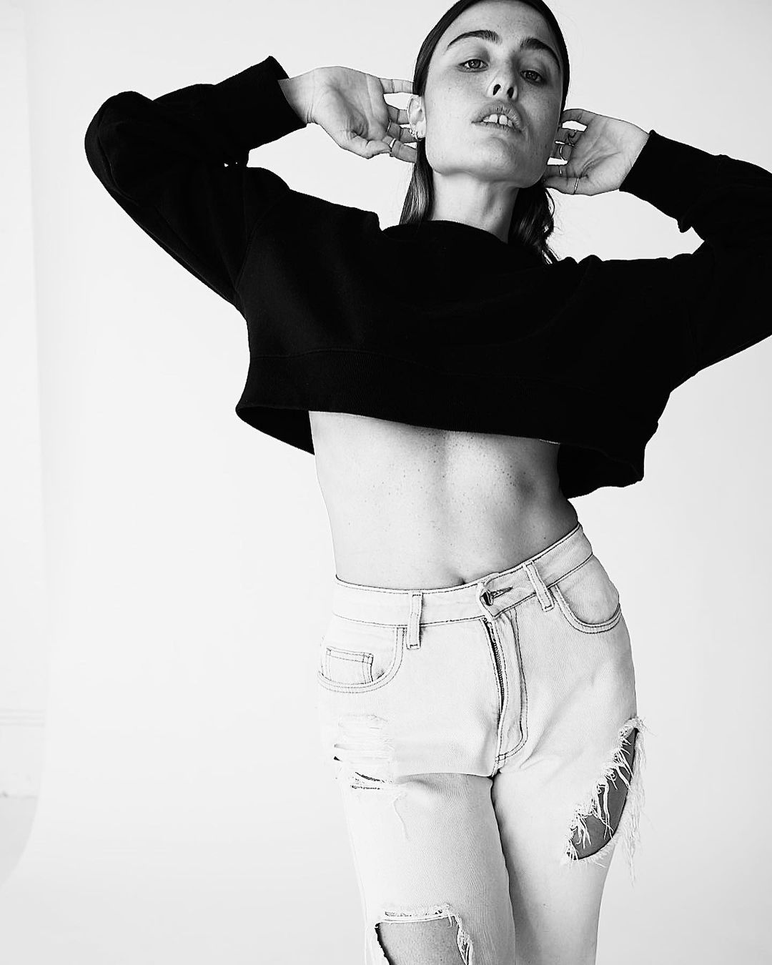 Monochrome portrait of a young woman in a black top and distressed light blue jeans against a plain background, posing with hands near head.