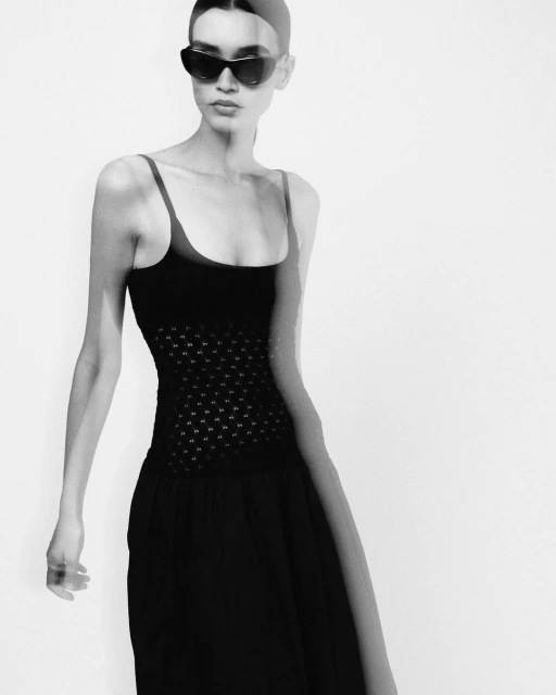 Black and white fashion portrait of a woman wearing a black dress and sunglasses against a bright background in a studio setting.