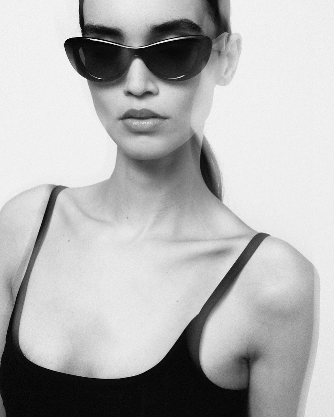 Close-up portrait of a woman wearing black sunglasses and a dark tank top against a plain white background by Daniel Nadel