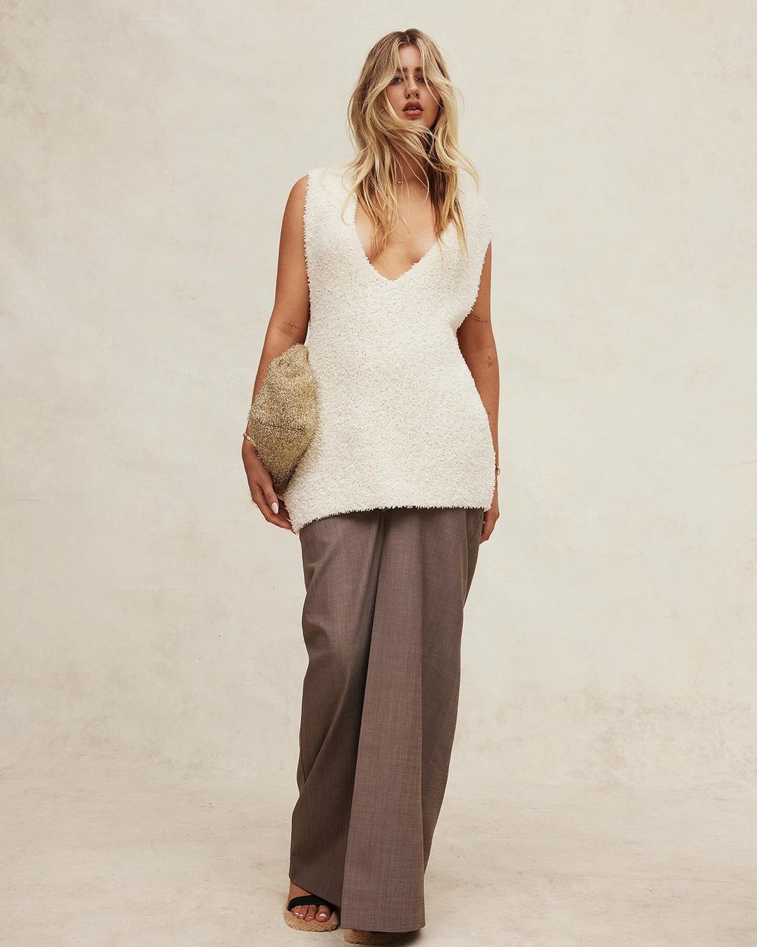 Full-length shot of an adult woman in an off-white sweater vest and wide-leg brown trousers, carrying a clutch against a plain backdrop.