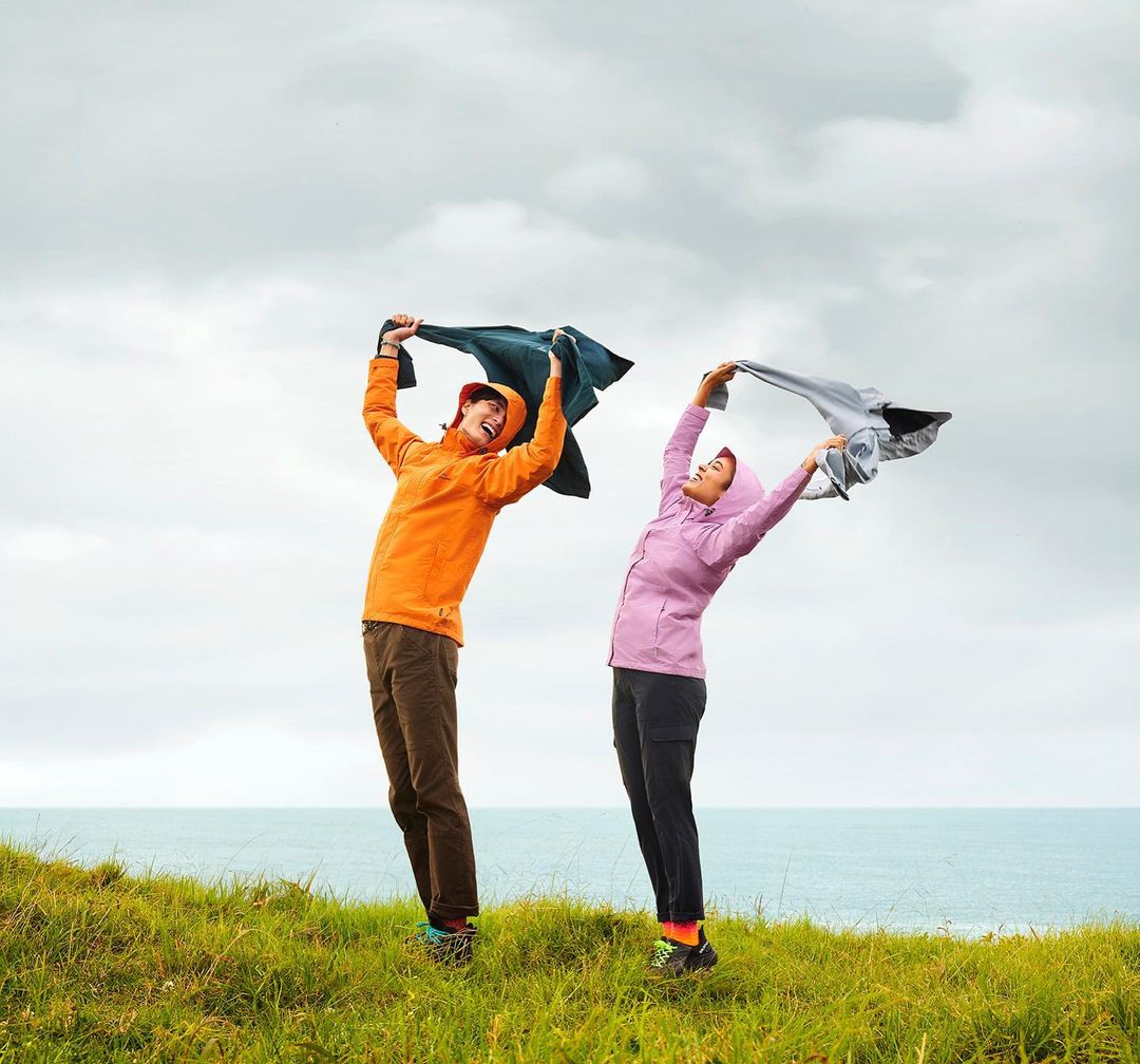 Two happy women in colorful rain jackets stand on a grassy hill by the ocean with overcast skies, holding jackets above their heads.