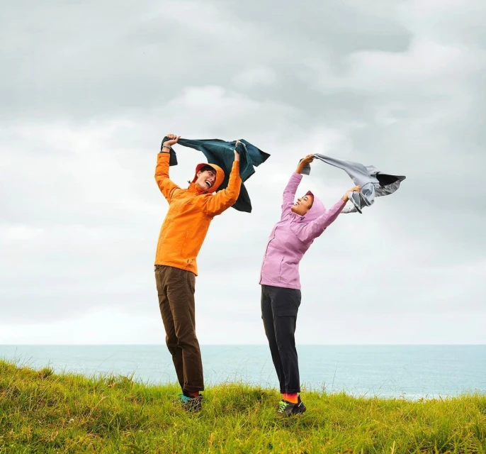 Two happy women in colorful rain jackets stand on a grassy hill by the ocean with overcast skies, holding jackets above their heads.