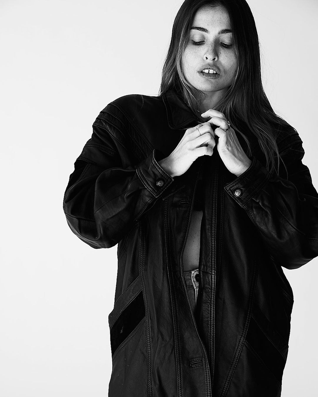 A woman with long brown hair buttons a vintage leather coat in a black and white studio portrait against a neutral backdrop.