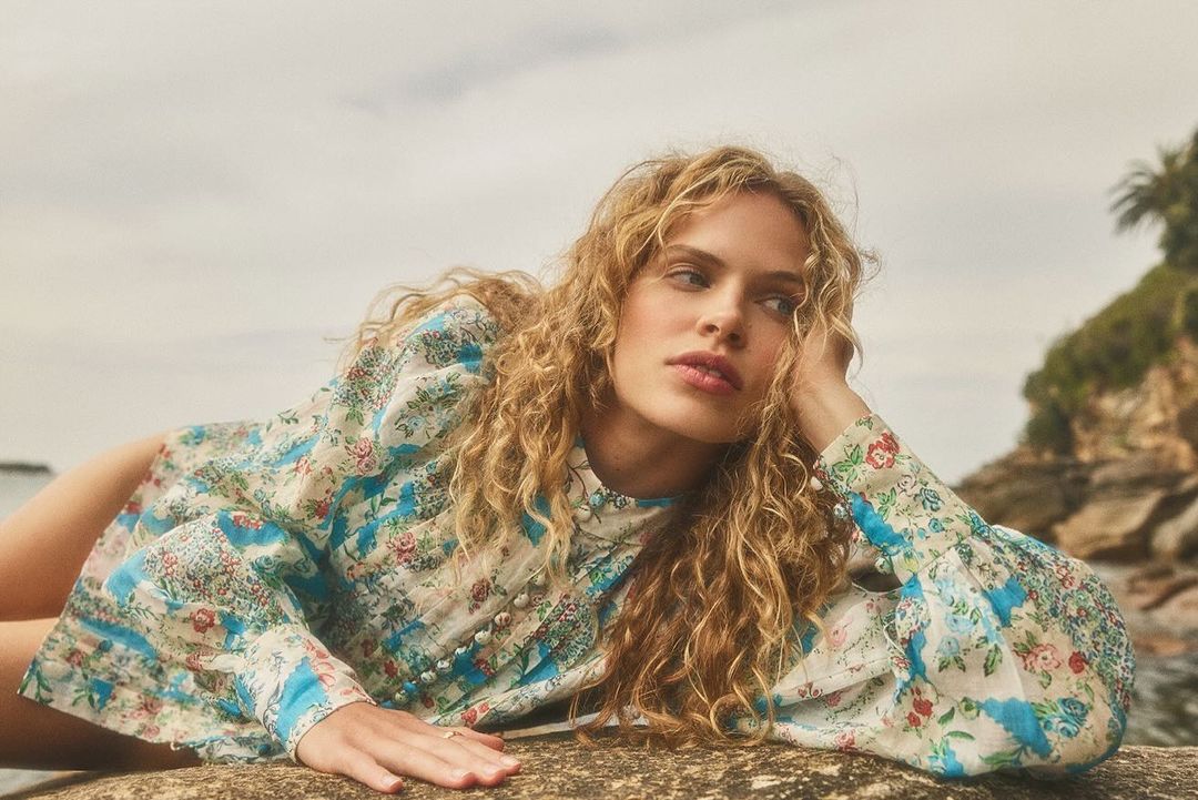 A young woman with curly blonde hair is reclining on a rock wearing a floral patterned blouse in an outdoor setting near the water.