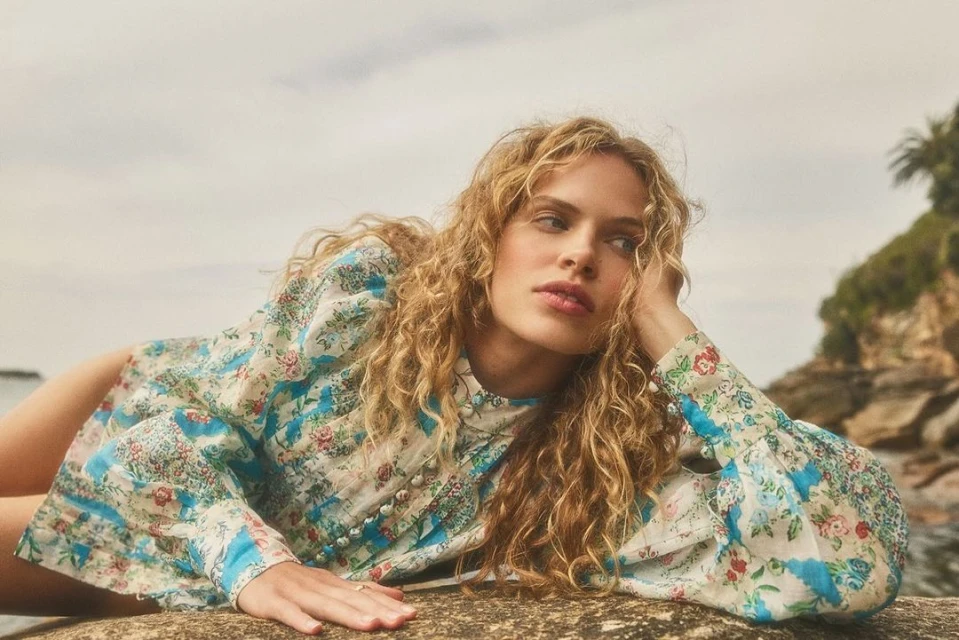 A young woman with curly blonde hair is reclining on a rock wearing a floral patterned blouse in an outdoor setting near the water.
