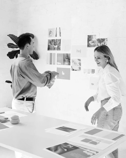 Two adults collaborate on design concepts in a bright studio. They review visual material pinned on a white wall during a meeting.