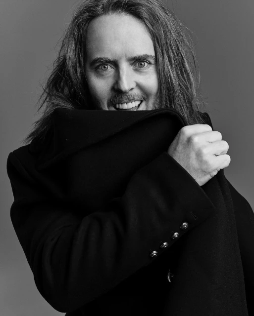 Black and white portrait of a smiling adult man with long hair, partially obscured by a black coat held up to his face indoors.