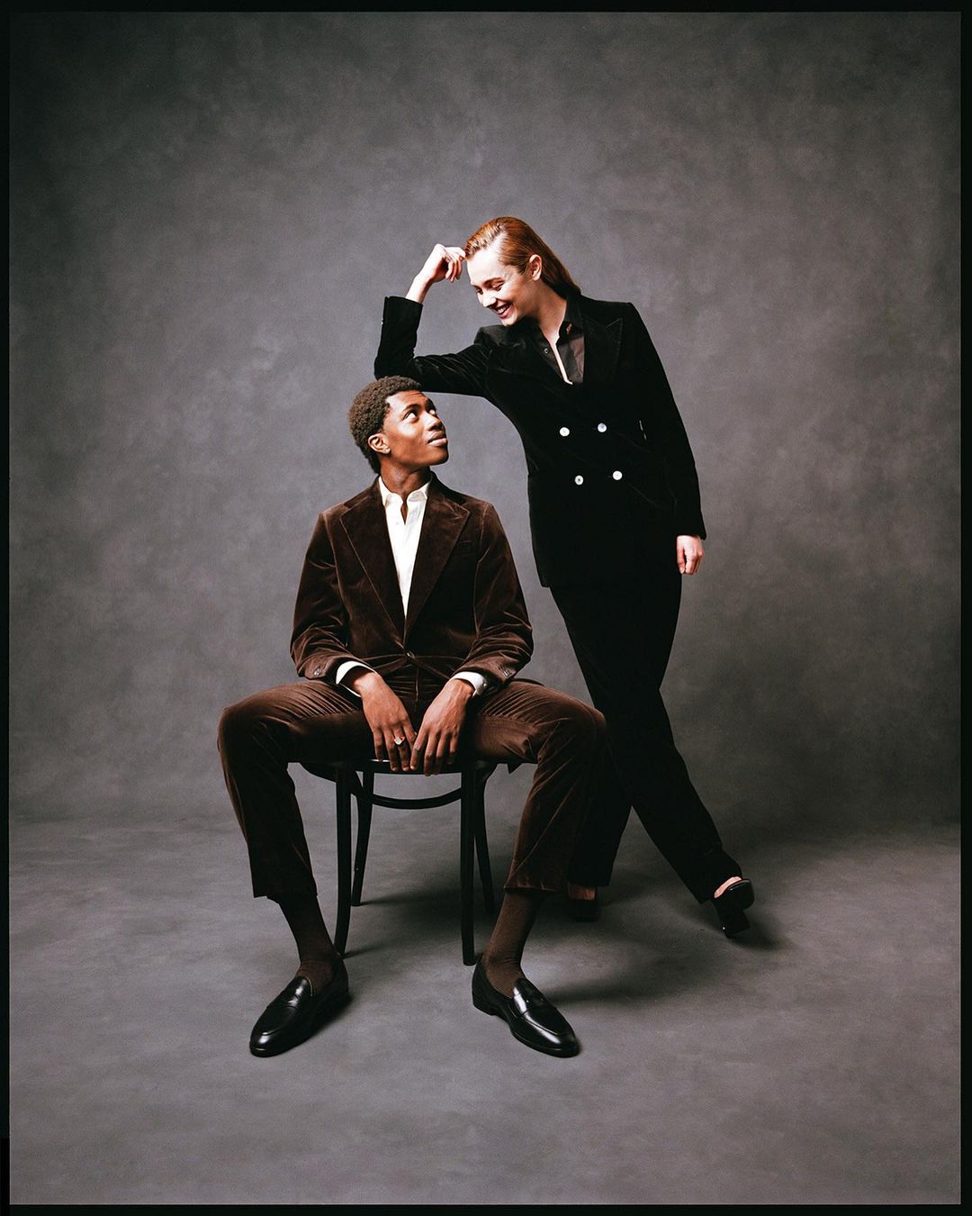 A stylish fashion portrait of an African American man sitting on a chair and a fair-skinned woman in black leaning on him in studio.