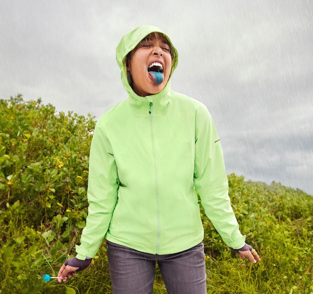 Woman in bright green hooded raincoat catches falling rain with blue tongue outdoors in nature. Overcast skies and landscape visible.