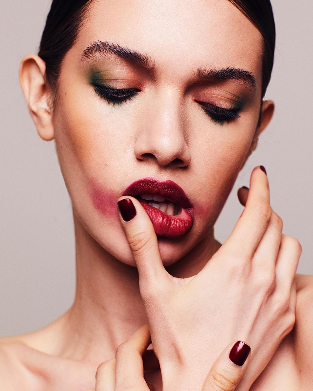 Close-up portrait of an adult woman with dramatic makeup, including bold red lipstick and green eyeshadow, posing with her finger in her mouth.