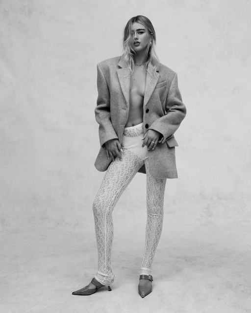 A slender fashion model poses in a gray wool blazer, sheer patterned leggings, and pointed-toe shoes against a neutral backdrop.