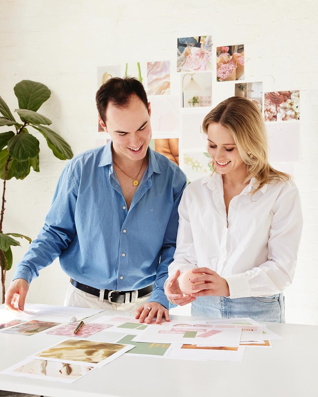 Two people, a man and a woman, standing at a table covered with papers, in an indoor setting, with white walls and a plant in the background.