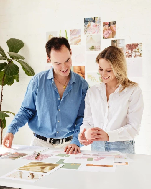 Two people, a man and a woman, standing at a table covered with papers, in an indoor setting, with white walls and a plant in the background.