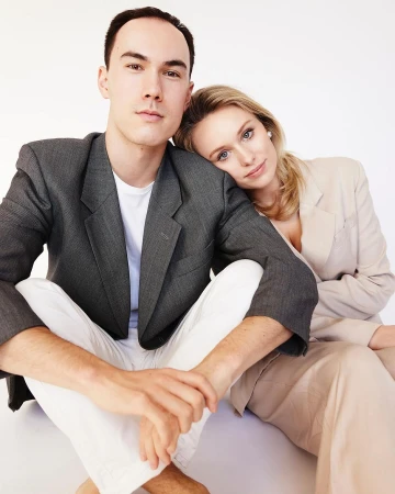 Man in grey blazer and white trousers seated next to a woman leaning her head on his shoulder against a plain white background.