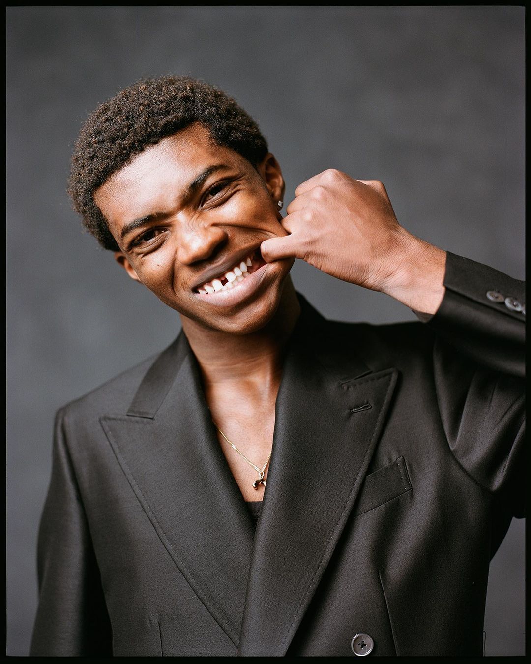 Portrait of a young African American man smiling with his finger pulling his cheek to show his teeth in front of a grey background.
