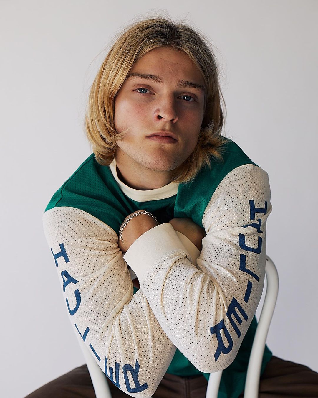 Blonde male model with green and cream long-sleeve shirt sitting on white chair in a close-up studio portrait with a neutral expression.