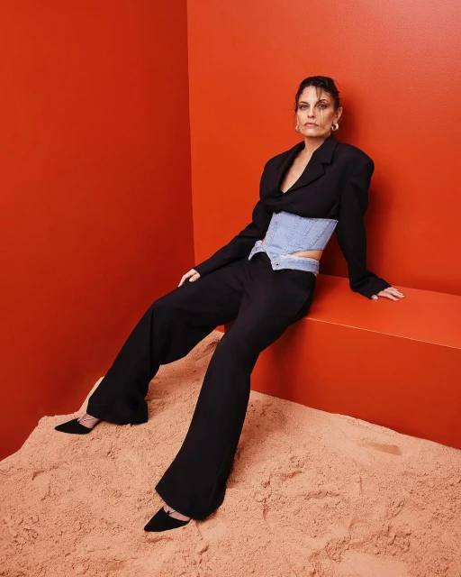 Fashionable woman in black suit and light blue corset poses on sandy surface against an orange backdrop in a studio setting.