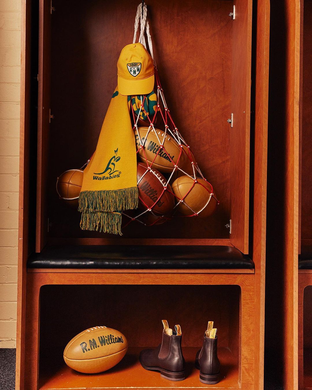 Locker room display with rugby balls, Wallabies scarf, cap, and boots arranged in wood locker with overhead and shelf storage.