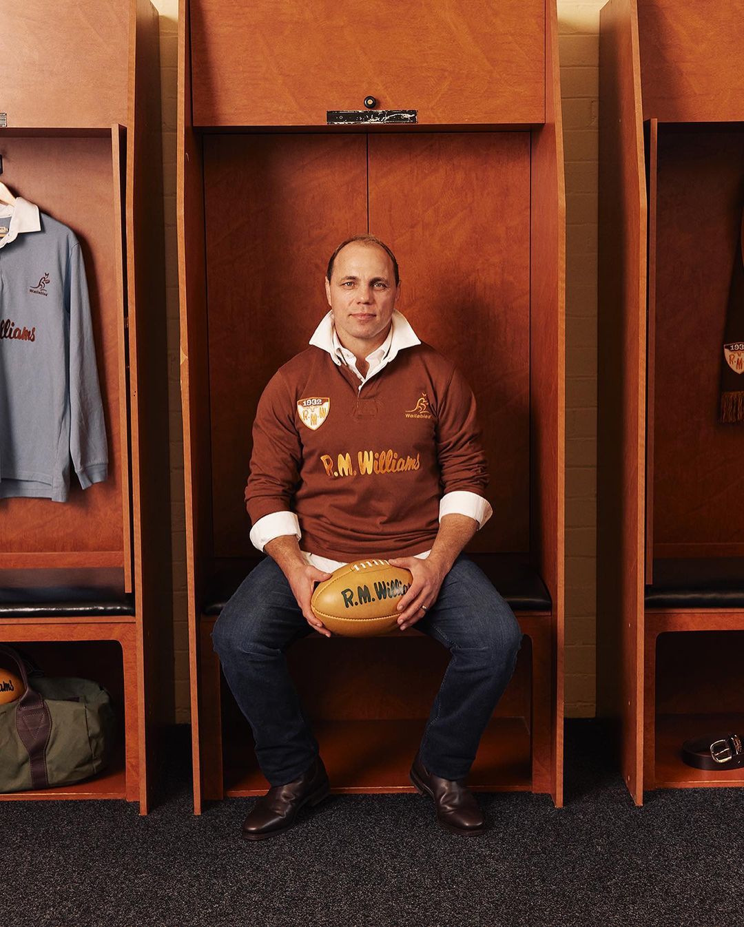 Man in brown rugby shirt holding an oval rugby ball, sitting in a wooden locker room with blue shirt visible. Interior shot, eye level.