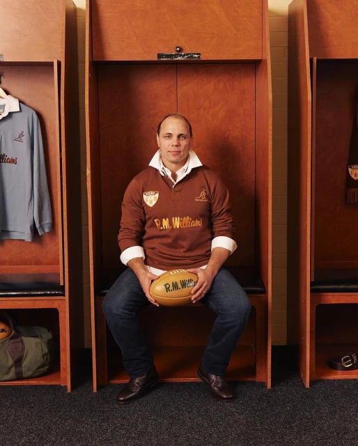 Man in brown rugby shirt holding an oval rugby ball, sitting in a wooden locker room with blue shirt visible. Interior shot, eye level.