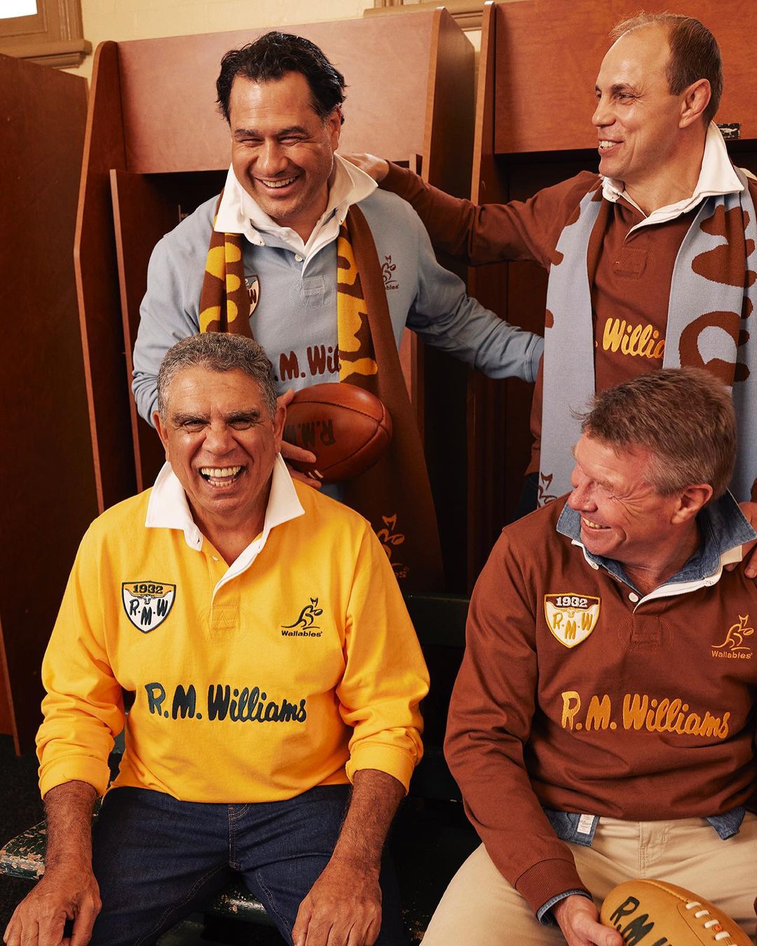 Four indigenous men smiling and wearing rugby shirts with R.M. Williams logos, posing together inside an indoor changing room.