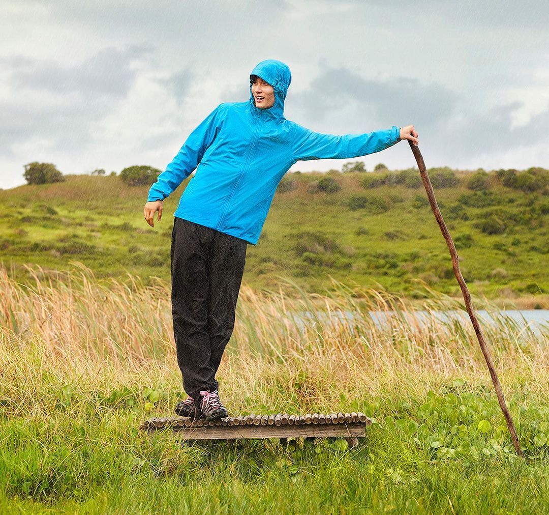 Adult woman in a blue hooded jacket, black pants and hiking boots poses with a wooden walking stick in a grassy rural environment.