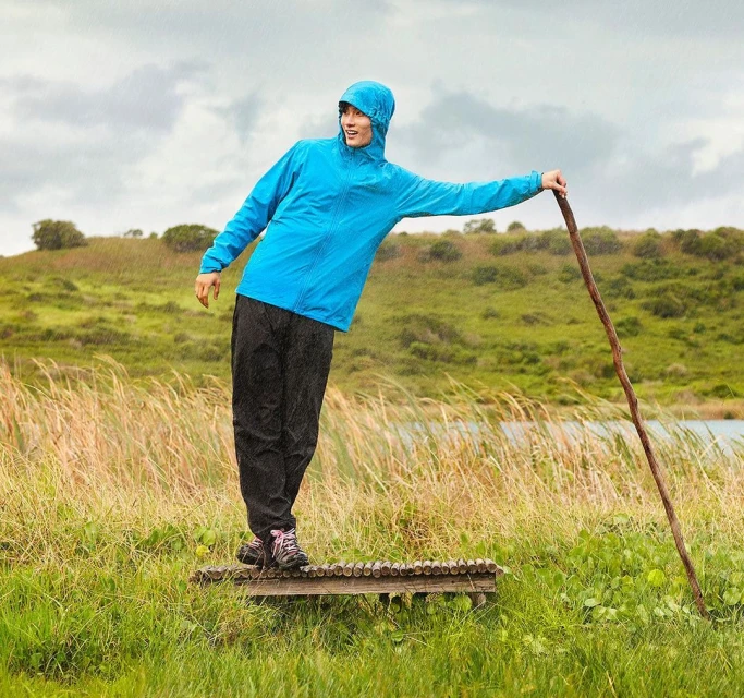Adult woman in a blue hooded jacket, black pants and hiking boots poses with a wooden walking stick in a grassy rural environment.