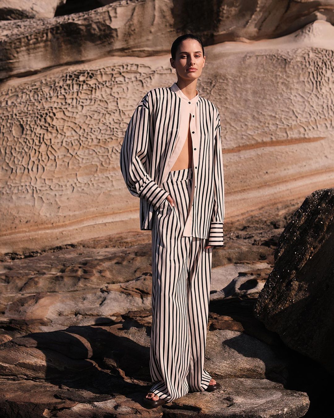 Fashion shot of a model in a striped pantsuit posing on a rocky beach, with a layered rock formation in the background.