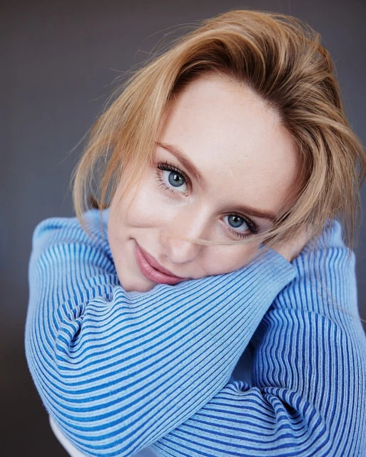 Blonde woman in blue sweater smiles at camera, resting head on arms indoors against grey background. Portrait, eye-level angle.