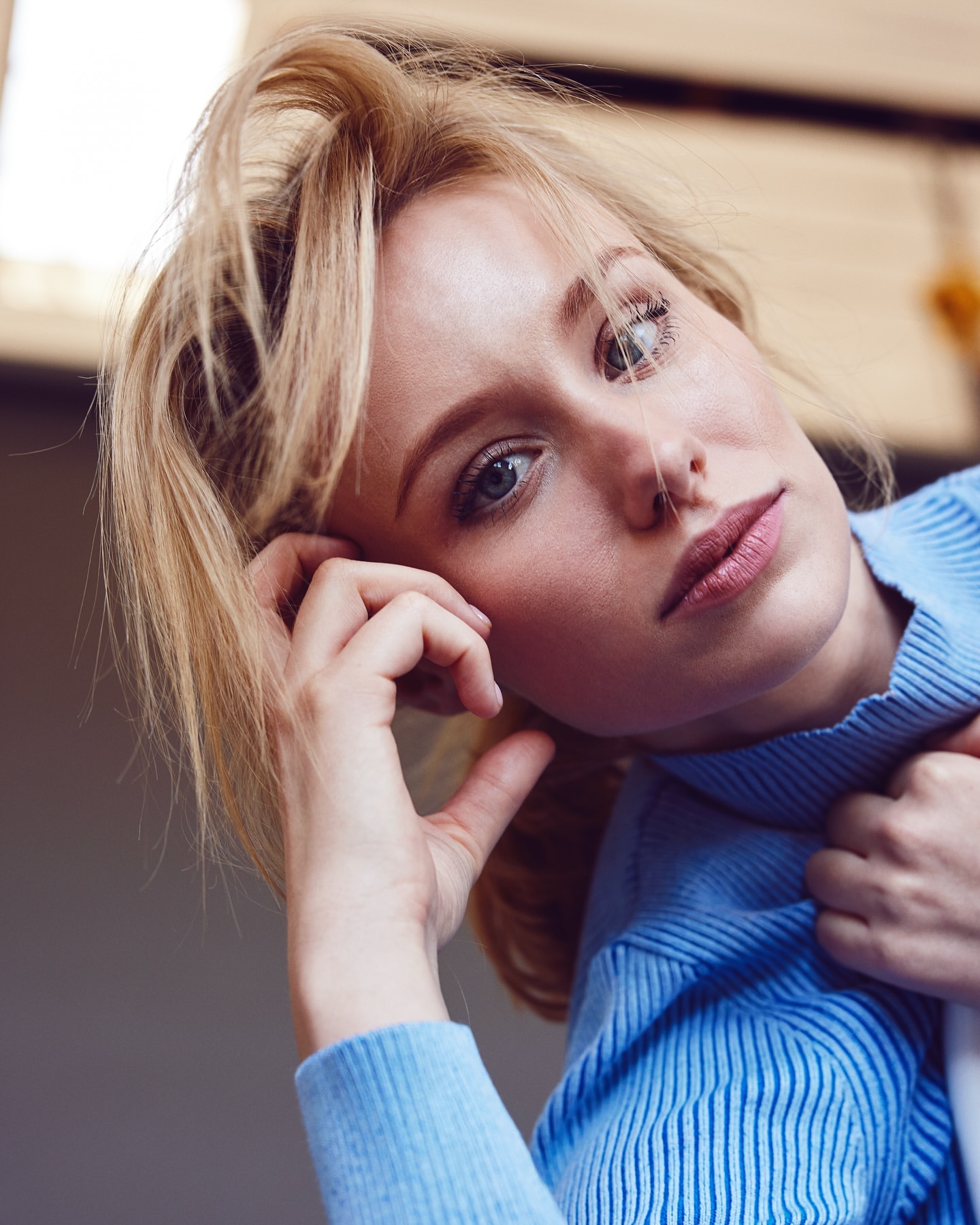 Close-up shot of an adult woman with blonde hair and blue eyes wearing a blue sweater, looking at the camera, indoors with natural lighting.