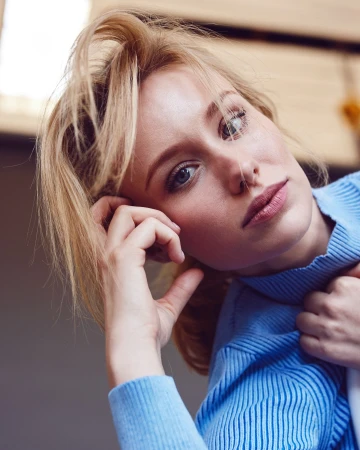 Close-up shot of an adult woman with blonde hair and blue eyes wearing a blue sweater, looking at the camera, indoors with natural lighting.