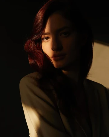 Portrait of a woman with red hair and light jacket illuminated by directional sunlight casting stark shadows across her face.