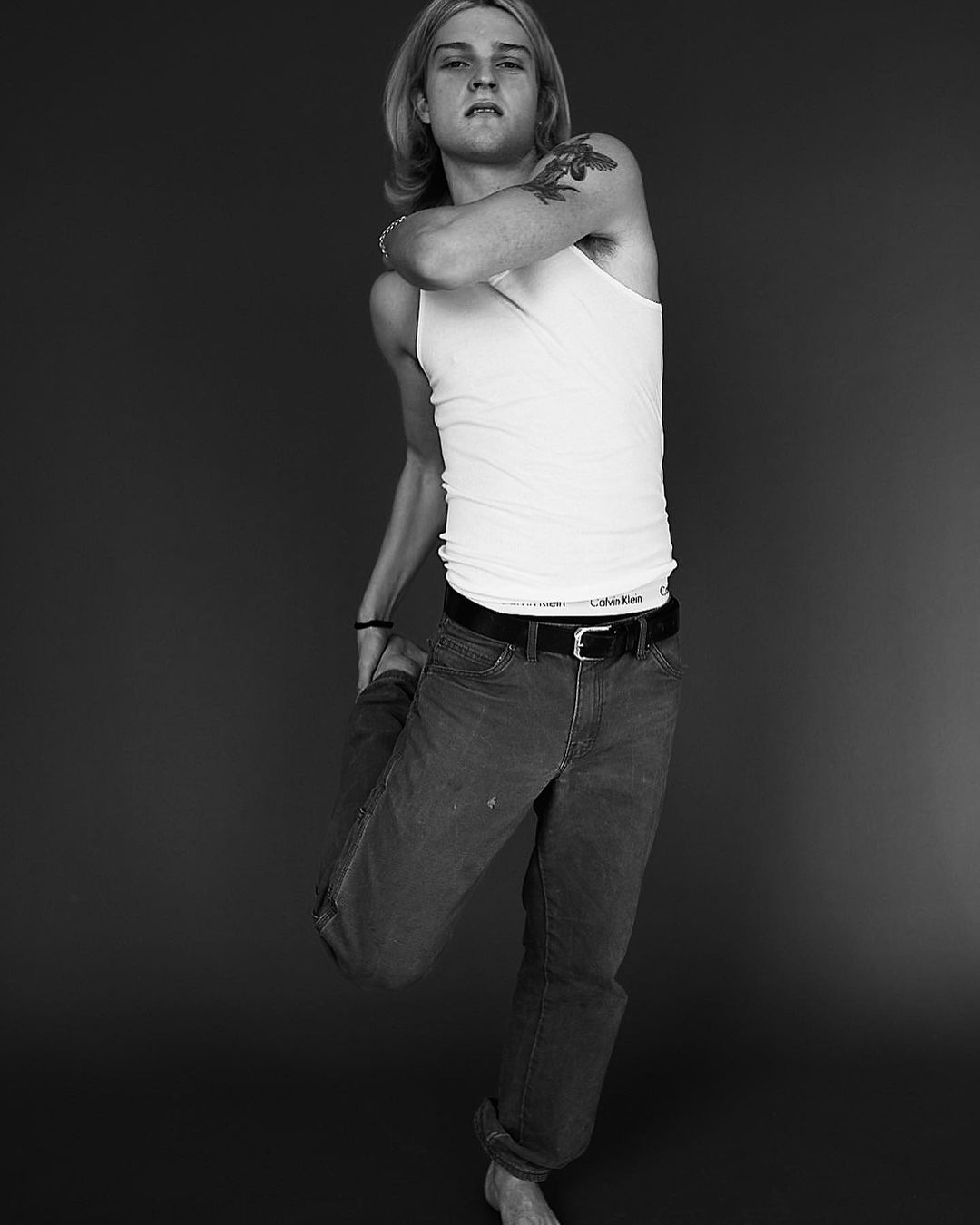 Black and white portrait of a young adult with long blond hair wearing a tank top, jeans, and bare feet, stretching a leg.