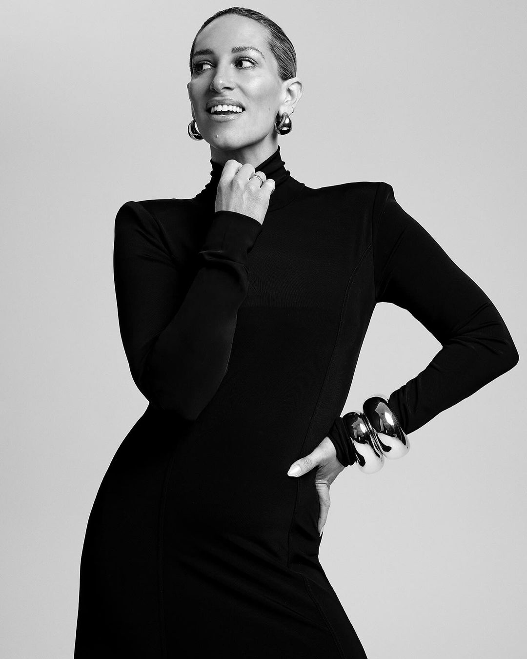 Black and white portrait of an adult female in a long-sleeved black turtleneck dress, posing with jewelry against a neutral backdrop.